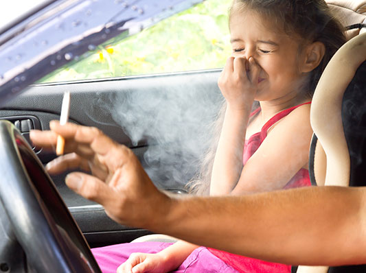 Little girl holding her nose and closing her eyes stting in the passenger seat while a male driving the car smokes