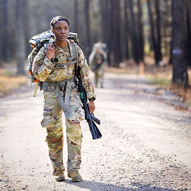 Service member with a pack and weapon walking along uneven ground