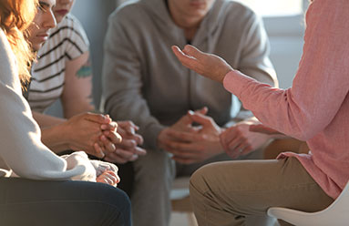 Support group. We see people sitting in a circle. Part of a face is visible, mostly hands.