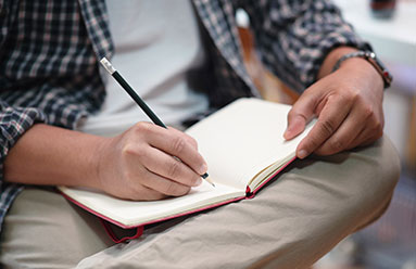 Image of a man visible from the waist down, writing in a journal that is in his lap