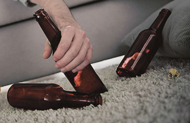 Empty beer bottles on the floor next to a couch. A man's arm hangs over indicating the man is unconcious