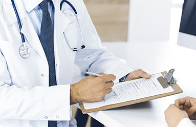 Cropped image of a doctor with a patient. We see the patient's hand as well as the doctor's torso. The doctor is holding a pen and a clipboard.